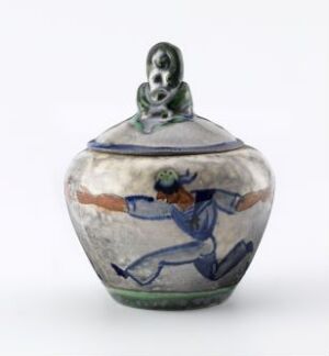  A small, decorative ceramic pot with a lid, featuring a dynamic, blue-painted figure in motion encircling the body and a simple human figure as the knob on the gray lid.