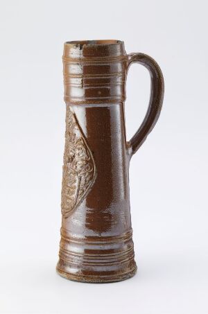  A tall, glossy chestnut brown ceramic beer stein with embossed decorations and ridged detailing on the upper and lower rims, featuring a handle on the side, against a neutral background. Artist name and title are unknown.