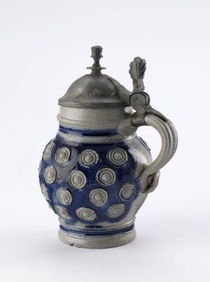  A decorative ceramic teapot with a deep cobalt blue glaze and white circular medallion patterns, adorned with a metal pewter lid featuring a stylized finial. The handle is designed with a rope-like motif, and the entire piece emanates a blend of functionality and ornate design.