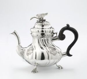  An elegant 'Tekanne' silver teapot by artist Daniel Erichsen. It features a lustrous, adorned body with embossed designs, a whimsical bird-shaped finial, and a curved, dark wooden handle.