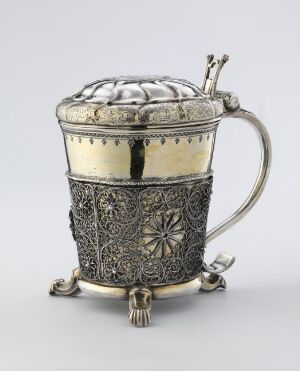  An ornate silver tankard with intricate filigree patterns and a decorated lid, featuring a curved handle and three small feet, sits against a light background.