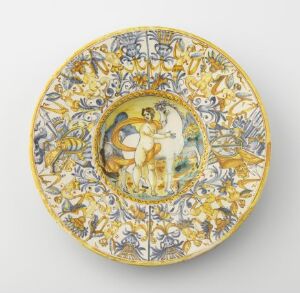  An ornate ceramic plate with scalloped edges, featuring a central medallion with classical figures in warm tones surrounded by a blue and golden-yellow floral and mythical motif. The glossy finish suggests fine craftsmanship.