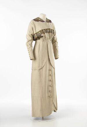  A full-length early 20th-century women's dress on a mannequin, featuring a muted beige color with dark brown trim on the collar and cuffs, a gathered waistband, decorative front buttons, and a structured skirt panel, set against a plain background. Artist Name and Title: Unknown.