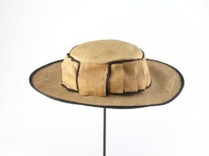  A wide-brimmed golden tan hat with a domed crown displayed on a black stand against a light grey background.