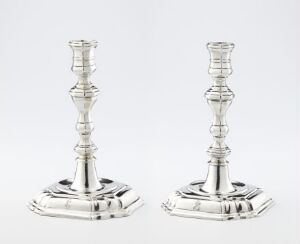  A pair of elegant, silver-colored metal candlesticks with intricate designs, featuring fluted stems and square bases, displayed against a neutral background.