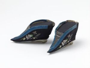  A pair of vintage, navy blue pointed shoes with an upward curve at the toe, adorned with delicate floral embroidery in light contrasting colors, and featuring a lighter blue band around the opening, set against an off-white background.