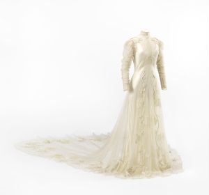  An elegant ivory dress with a high collar, long sheer sleeves, and an extended trailing train adorned with floral patterns on a mannequin against a plain light background, suggesting a vintage bridal gown.