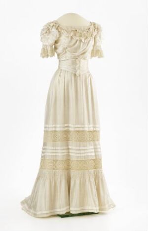  A vintage-style cream-colored dress with horizontal beige bands and floral embellishments on the bodice and shoulders, displayed against a plain background.