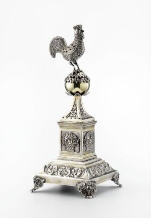  An ornate, silver, rooster-shaped sculpture on a tiered base with decorative floral patterns and clawed feet, indicative of luxurious craftsmanship. Artist name and title unknown.