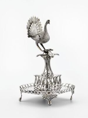  An intricately crafted silver peacock sculpture perched elegantly on a decorative pedestal with a lacy, circular base against a white background.