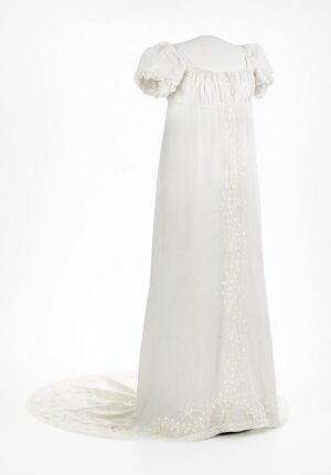  An elegant, long white dress with a fitted bodice featuring vertical embroidered panels, short puffed sleeves with lace details, and a flowing skirt extending into a lengthy train, displayed against a white backdrop.