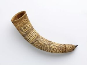  An intricately carved, curved, horn-like object with light to mid-brown tones set against a plain white background. The surface features detailed patterns or symbols, reminiscent of an artifact. The artist's name and title are unknown.