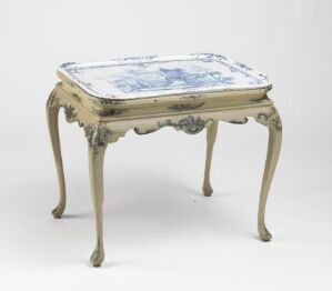  A vintage-style table with ornate, curvy legs and a frame in muted grayish-beige tones, featuring a tabletop with a delft blue and white tile tableau in a pastoral or mythological scene.