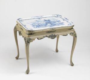  An ornate table with curved legs and decorative carvings, painted in a creamy white color that has a distressed finish. The tabletop features a recessed blue and white illustration with a pastoral scene, resembling Delft porcelain designs.