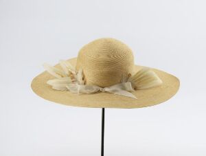  A wide-brimmed beige hat with a smooth crown and a light-colored band tied in a bow, displayed on a black stand against a light-gray background.