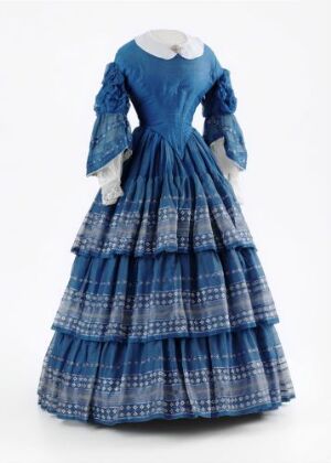  An antique blue and white tiered dress with full sleeves and a small white collar, displayed against a light grey background. The dress exhibits mid-Victorian design, featuring rich cobalt blue fabric for the bodice and a patterned skirt with horizontal bands of white and light blue. The layered skirt suggests the form was supported by a structural undergarment typical of the era.