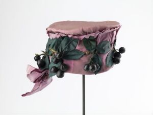  A vintage-style women's hat with a ruffled dusky pink brim, adorned with a garland of deep green leaves and black berries, displayed on a stand against a white background.