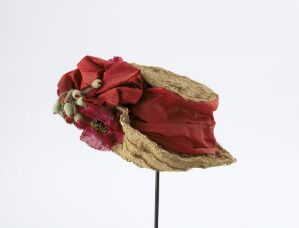  A vintage-style straw hat with a faded red fabric band and decorative beads and flowers on the side, displayed on a dark metal stand against a light grey background.