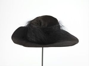  A black fashion hat with a broad brim and a band of wispy hair-like material, displayed on a thin stand against a white background.