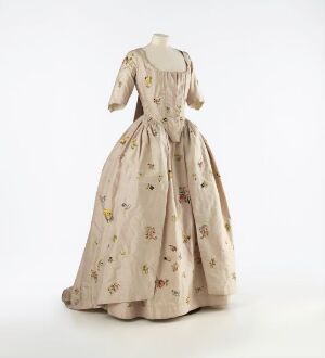  A historical dress from the 18th or early 19th century in a pale cream color with soft pastel floral motifs, featuring a full skirt, fitted bodice, and short puffed sleeves, displayed against a light background.