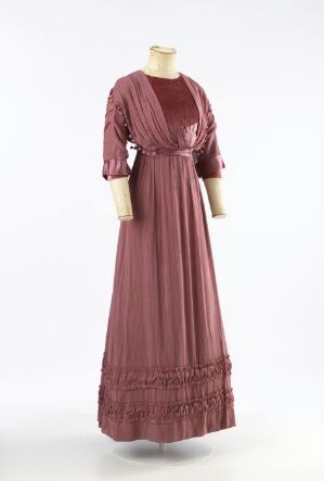  A vintage dusty rose-colored dress with long puffed sleeves, a round neckline, and a ruffled hem on a mannequin against a neutral background.