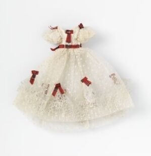  A miniature, historically-styled doll's dress in an off-white color with dark red accents, including bows at the collar, waist, and on the full, layered skirt, displayed against a light gray background. Artist Name and Title are unknown.