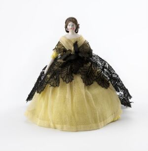 
 A historical figurine of a woman dressed in a pale yellow gown with a black lace shawl, against a plain light background. The figurine's hair is styled in an updo, and her facial features are delicately outlined. Artist name and title are unknown.