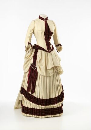  A late Victorian-era styled dress displayed on a headless and armless mannequin. The dress features a light beige bodice with a subtle stripe pattern and a high ruffled neckline adorned with a burgundy ribbon. The skirt is full and floor-length, embellished with wide burgundy velvet stripes and smaller striped detailing near the hem. The dress suggests the formal fashion of the late 19th century.