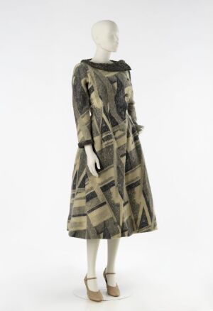  A mannequin wearing a mid-calf length vintage dress with long sleeves, patterned with abstract geometric shapes in shades of brown and muted green, against an off-white background.