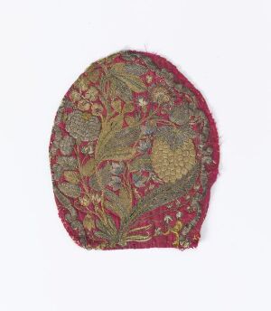  An oval-shaped piece of textile with an ornate botanical design in muted shades of red, green, yellow, and brown, potentially a segment of historical headwear or a decorative fabric panel.