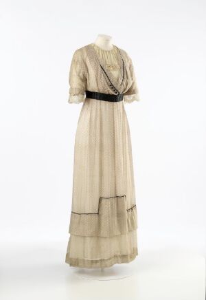  A historical ivory-colored dress with full-length sheer sleeves and a draped bodice, cinched at the waist with a black belt, against a plain background.