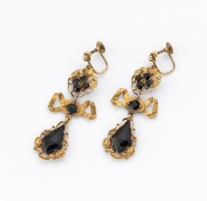  A pair of ornate gold-toned earrings with black jewels and teardrop-shaped stones, featuring intricate scroll detailing and a cross-like mid-section, showcased against a light background.