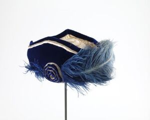  An antique-looking navy blue ceremonial hat with gold trim, a spiraled decoration on the side, and a large plume of blue feathers, displayed on a stand against a light background. Artist name and title are unknown.