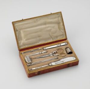  Antique surgical instrument kit composed of polished metal tools, including a scalpel, scissors, and various probes, displayed on a maroon velvet lining inside an open, brown leather box with creamy beige silk lining.