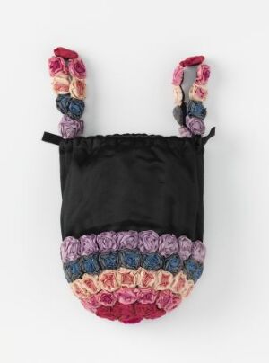  A decorative handcrafted bag featuring a solid black upper section and a multicolored, textured lower trim. The straps are adorned with fabric flowers in pink, red, blue, and violet shades, with each strap tied with a black bow.