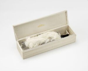  An open, light beige rectangular box with a hinged lid, lined with a similar fabric, containing delicate white handmade textile objects, possibly lace, arranged neatly inside, on a white neutral background.