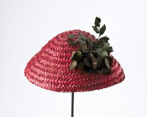  A crimson red woven hat with a broad, downward-sloping brim and a cluster of dark green leaves as decoration, displayed against a pale grey background on a vertical stand.